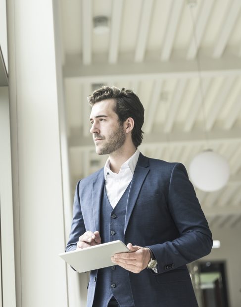 Successful manager standing in modern office building, using laptop, looking out of window